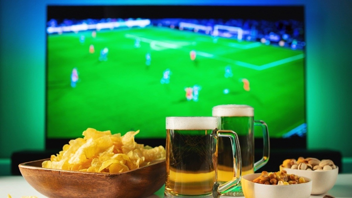 Domácí sledování fotbalu s pivem, chipsy a ambientním osvětlením za televizí